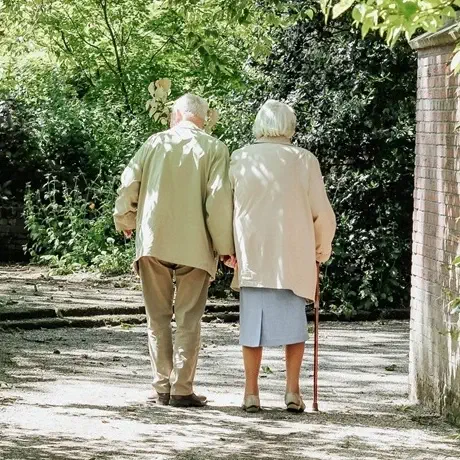 Elderly couple walking hand in hand down a sunlit garden path.